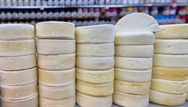 Taze peynir tekerleklerinin yakın plan görüntüsü düzgün istiflenmiş, kremsi dokusunu ve zengin rengini bir pazar ortamında sergiliyor. Arkaplanda paketlenmiş ürünlerle dolu raflar bulanıklaştırılıyor.