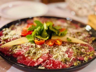 A beautifully served beef carpaccio garnished with fresh arugula, cherry tomatoes, parmesan shavings and drizzled with olive oil and pesto sauce on an elegant black plate. clipart