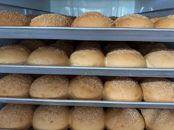Taze pişmiş hamburgerlerle dolu üç metal tepsi. Susam tohumlarıyla kaplı. Hepsi de fırın ortamında eşit olarak yerleştirilmiş..