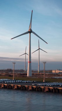 Emden, Germany - 02 04 2023: Parking place with the brand new vehicles in the area of the biggest german car manufacturer with huge wind generators in the background