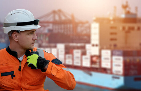 Offshore technician. Seafarer. Seaman. Navigator. A man in a boiler suit wearing a safety helmet with a container ship in the background. Seafarer in front of the container ship with a radio station.