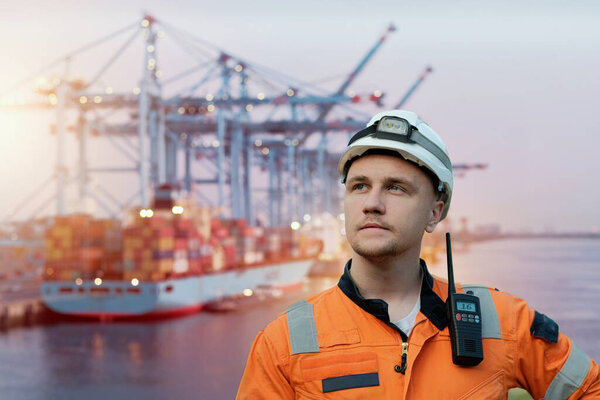Wind Farm Offshore Maintenance Technician. Seafarer. Seaman. Navigator. A Man In A Working Overall Boiler Suit With A Radio And Safety Helmet With A Blurred Container Vessel In The Background