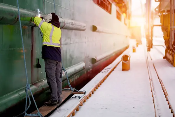 Certified Welder Technician Wearing Protective Clothes And Equipment While Standing On A Secured Wooden Step And Working With A Welding Machine To Repair The Crack On The Hull Of The Merchant Vessel