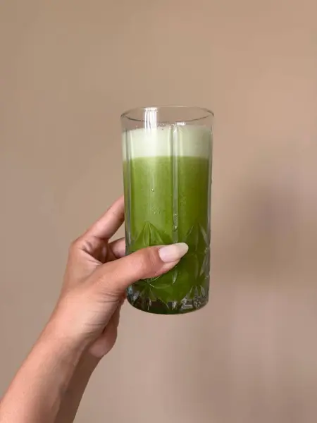 Uzun ve berrak beyaz tırnakları olan bir el kum rengi arka planda bir bardak yeşil detoks suyu tutuyor. 