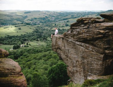 Curbar Edge 'de vadiye bakan bir kayanın üzerinde oturan bir adam. Sahne huzurlu ve sakin, manzaranın ve bölgenin sessizliğinin tadını çıkaran bir adam.