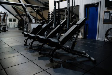 Spor salonunda dizilmiş üç siyah egzersiz bankı var. Banklar boş ve spor salonu loş bir ışık altında.