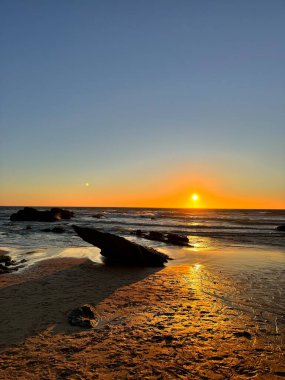 Kumlu bir sahili ve ön planda keskin bir kayası olan çarpıcı bir okyanus kıyısı. Gökyüzü gün batımında canlı turuncu ve kırmızı renklerle parlayarak Portekiz kıyılarının güzelliğini yakalıyor..