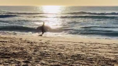 Günbatımında okyanusa doğru koşan sörfçü Portekiz 'de altın bir günbatımında sahile doğru koşan bir sörfçünün silüeti. Açık hava macerası ve özgürlük için mükemmel bir an..