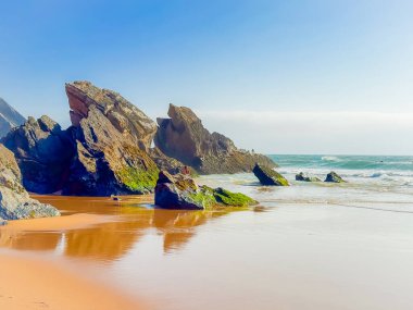 Uzun ve dramatik bir uçurumla çevrili kumlu bir sahil. Kaya ve okyanus arasında güçlü dikey kontrastlar