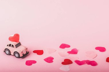  A small pink car model carries heart-shaped marmalade on a pink background among paper hearts. Concept for Valentine's Day.