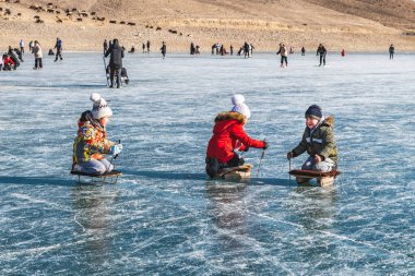 Lake Ara-Kol, Naryn region, Kyrgyzstan - January 5, 2024: Boys and girls have fun and ride on homemade sleds on the frozen lake. Activity during the winter holidays. clipart