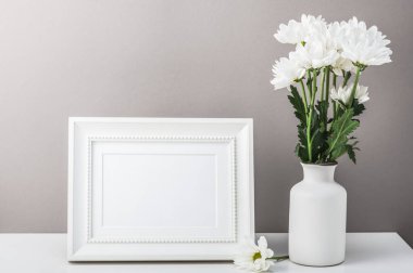 Boş bir fotoğraf çerçevesi ve bir buket beyaz kasımpatı, gri arka planda beyaz bir vazoda papatyalar. Kartpostal, tebrik, ev içi..