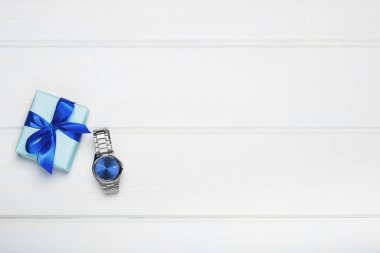 Mavi fiyonklu bir hediye kutusu ve beyaz ahşap arka planda mavi kadranlı bir erkek saati. Babalar gününü, oğulların gününü, yeni yılı kutluyoruz..