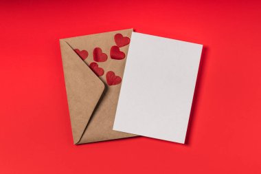 Kraft kağıt zarf, boş form ve kırmızı arka planda kağıt kalp konfetisi. Sevgililer Günü veya doğum günü tebrik kartı maketi.