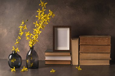 Çiçek açan sarı forsythia dalları, boş fotoğraf çerçevesi ve kahverengi arka planda bir yığın kitabı olan iki cam vazo. Bahar çiçeği arkaplanı.