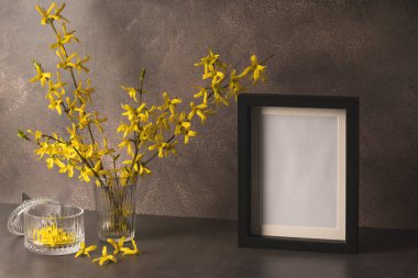 Sarı forsythia dalları açan bir bardak ve kahverengi arka planda boş bir fotoğraf çerçevesi. Bahar çiçeği arkaplanı