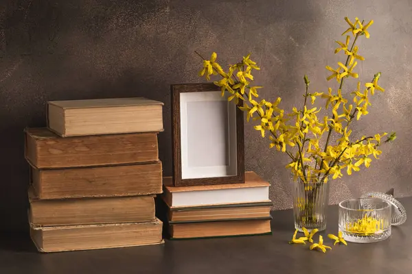 Çiçek açan sarı forsythia dalları boş bir fotoğraf çerçevesi ve kahverengi arka planda bir yığın kitap olan bir bardak. Bahar çiçeği arkaplanı.