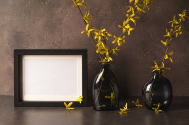 Forsythia dalları ve kahverengi arkaplanda boş fotoğraf çerçevesi olan iki şık cam vazo..