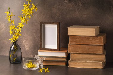 Kitap yığınları, boş fotoğraf çerçevesi ve kahverengi arka planda çiçek açan vazolar..
