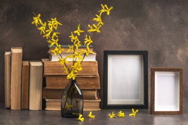 Süslü cam vazo, çiçek açan fındık dalları, kitap yığınları ve kahverengi arka planda iki boş fotoğraf çerçevesi..