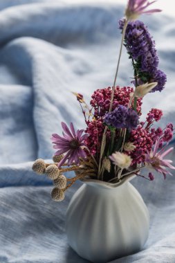 Keten açık mavi masa örtüsünde kurutulmuş çiçeklerle seramik vazo. Bahar narin çiçekli arka plan, seçici odak.