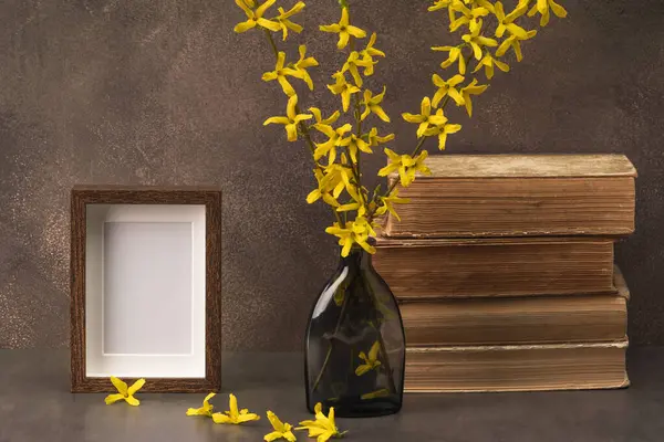 Süslü cam vazo, çiçek açan çanak çömlek dalları, kitap yığınları ve kahverengi zemin üzerinde boş fotoğraf çerçevesi.