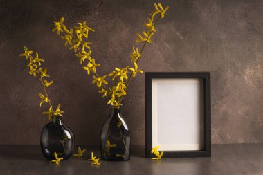 İki şık cam vazo çiçek açan fındık çiçekleri ve kahverengi bir arka planda boş bir fotoğraf çerçevesi. Çiçek dekoru..