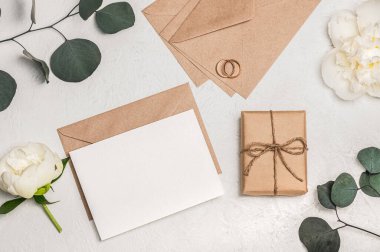 Düğün davetiyesi ya da tebrik kartı modeli el işi zarf, hediye kutusu, beyaz şakayık çiçekleri ve okaliptüs dalları ve arka planda alyanslar..
