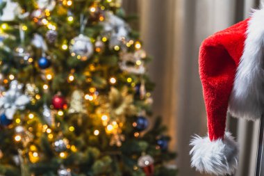 Noel Baba şapkası TV 'nin köşesinde arka planda asılı. Işıklı çelenklerle bir Noel ağacının arkasında. Bulanık arkaplan, seçici odak.