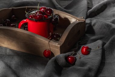 Ahşap bir tepsi ve gri bir masa örtüsünde olgun kirazlarla dolu kırmızı bir kupa..