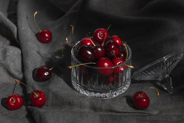 Gri bir masa örtüsünde olgun kırmızı kirazlı cam bir vazo..