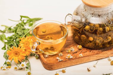 Çaydanlıkta taze demlenmiş papatya çayı, bir fincan çiçek çayı ve beyaz bir masada şifalı otlar. Organik doğal ilaç. Seçici odaklanma..