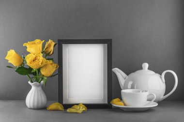 Boş fotoğraf çerçevesi, seramik vazoda sarı güller, beyaz çaydanlık ve gri arkaplanda bir fincan çay..