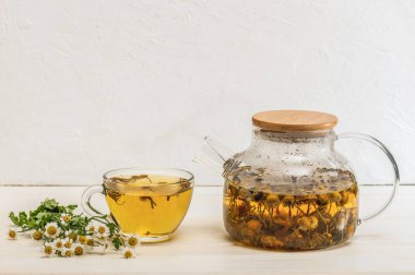 Çaydanlıkta taze demlenmiş papatya çayı, bir fincan çiçek çayı ve beyaz bir masada papatya çiçeği. Organik doğal ilaç. Seçici odaklanma..