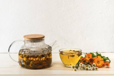 Cam çaydanlıkta tıbbi papatya çayı, bir fincan çay ve papatya çayı ve beyaz bir masada takvim çiçeği. Organik doğal tıp.