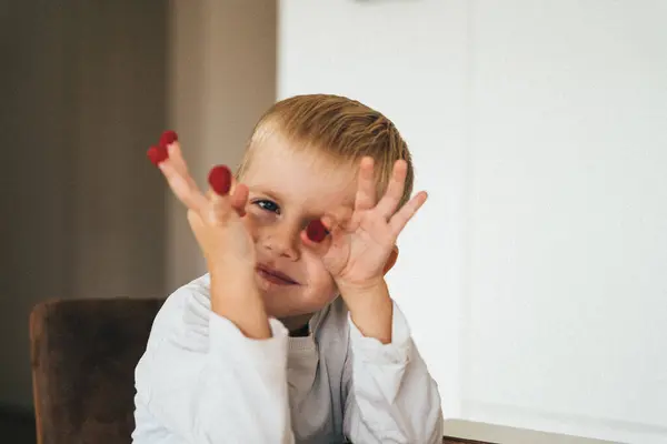 smiling boy of 4 years old sitting in the kitchen at the table with a raspberry on each finger . High quality photo