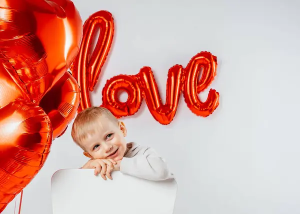 A blond boy sits on a chair surrounded by heart-shaped balloons with the word Love written on the wall. The scene exudes a warm, festive Valentines Day vibe, combining innocence and celebration in a