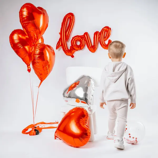 A blond boy sits on a chair surrounded by heart-shaped balloons with the word Love written on the wall. The scene exudes a warm, festive Valentines Day vibe, combining innocence and celebration in a