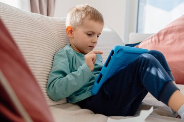 4 yaşındaki bir çocuk elinde tabletle evdeki kanepede oturuyor. Yüksek kalite fotoğraf