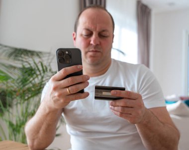 Elinde kredi kartı ve cep telefonu olan bir adam evdeki oturma odasında bir sandalyeye oturur. Yüksek kalite fotoğraf