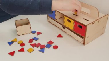 Küçük bir öğrenci, göğsünde çok renkli kutular bulunan çok renkli geometrik şekiller düzenler: kırmızı, sarı, yeşil ve mavi. Masanın üzerine farklı renklerin geometrik şekilleri saçılmış.. 