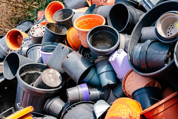 A large pile of used plastic plant pots in black, orange, and purple shades, stacked haphazardly outdoors. The worn and dirty containers highlight themes of recycling, waste management, and gardening