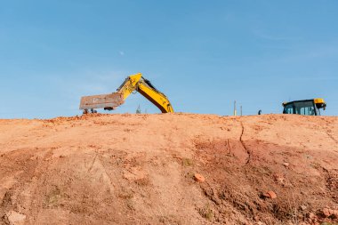Sarı kazıcı açık mavi gökyüzüne karşı bir inşaat sahasında toprağı kazıyor. Makinelerin kolu toprak tümseğin kenarına kadar uzanıyor ve inşaat faaliyetlerini gösteriyor.