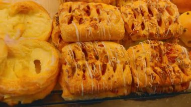 Fırın vitrininde sergilenen çeşitli altın sarısı, taze pişmiş çörekler. Yumuşak üzümlü çörekler ve yumuşak kabuklu gevrek reçelli börekler pasta severler için davetkar ve lezzetli bir seçim oluşturur.