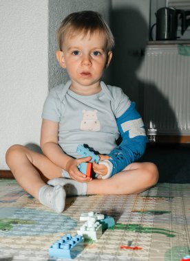 Yerde oturan kolları alçılı, renkli yapı taşlarıyla meşgul genç bir çocuk. Oyun zamanı oyuncaklarla uğraşırken odaklanmış ifade. Yaratıcılık ve esneklik sahnesi