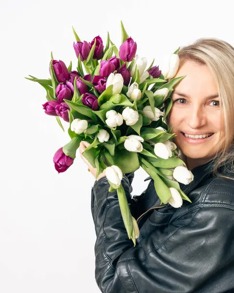 Siyah deri ceketli neşeli sarışın bir kadın büyük bir buket beyaz ve mor lale tutuyor. Çiçeklere bakarken gülümsüyor, temiz beyaz bir arkaplan yaratıyor.