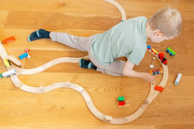 Küçük çocuk ahşap bir zeminde renkli bir oyuncak trenle dikkatle oynuyor. Çocuk raylara ve trenlere bağlanıyor, yaratıcılığını ve motor becerilerini geliştiriyor.