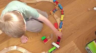 Dört yaşında neşeli bir çocuk yerde tahta bir oyuncak trenle oynuyor. Yukarıdan aşağı görünüm, hayal gücünü, odağı ve motor becerileri geliştiren renkli tren ve rayları gösterir.
