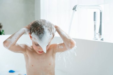 Çocuk küvette saç yıkıyor, sabunlu köpüklerle kaplı, musluktan akan su. Sahne banyo zamanındaki neşeli ve eğlenceli anı yakalıyor, temizlik ve eğlenceyi vurguluyor.. 