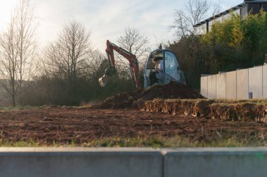 İnşaat alanında kovayla kil ve kayalık toprak temizleyen bir kazıcı. Kalkınma ve mühendisliği sembolize eden ağır makineler, yerleşim alanında iş yapmak için zemin hazırlıyor